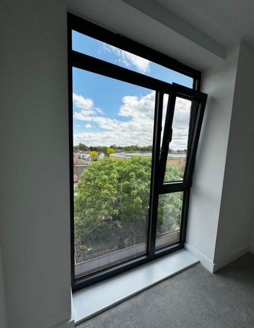 Black aluminium tilt-and-turn window tilted open above a tree-lined street, seen from inside a finished room