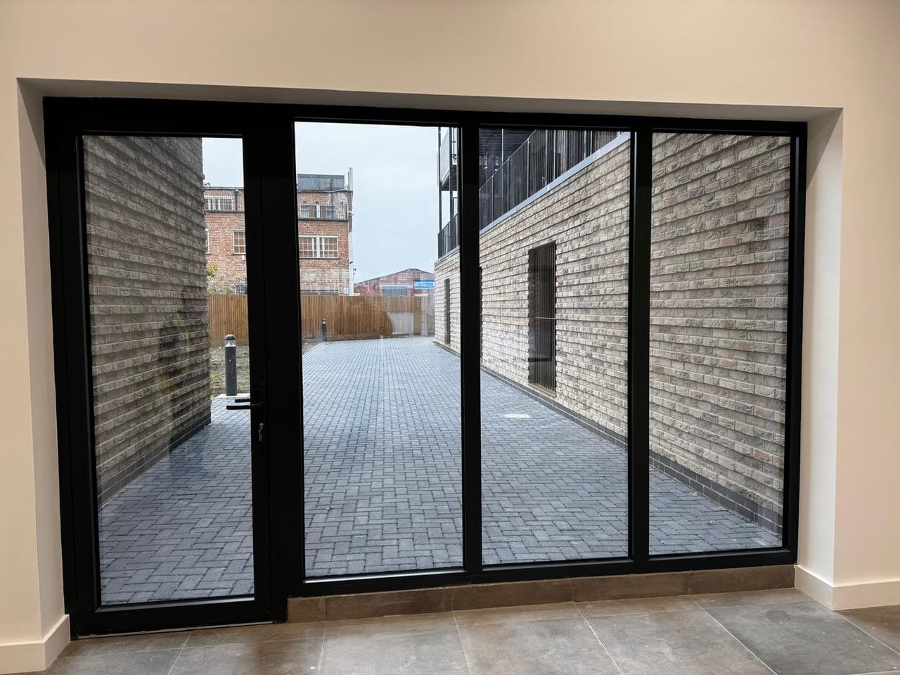 Wide aluminium door set with fixed side panes seen from inside a ground-floor apartment, opening onto a block-paved courtyard